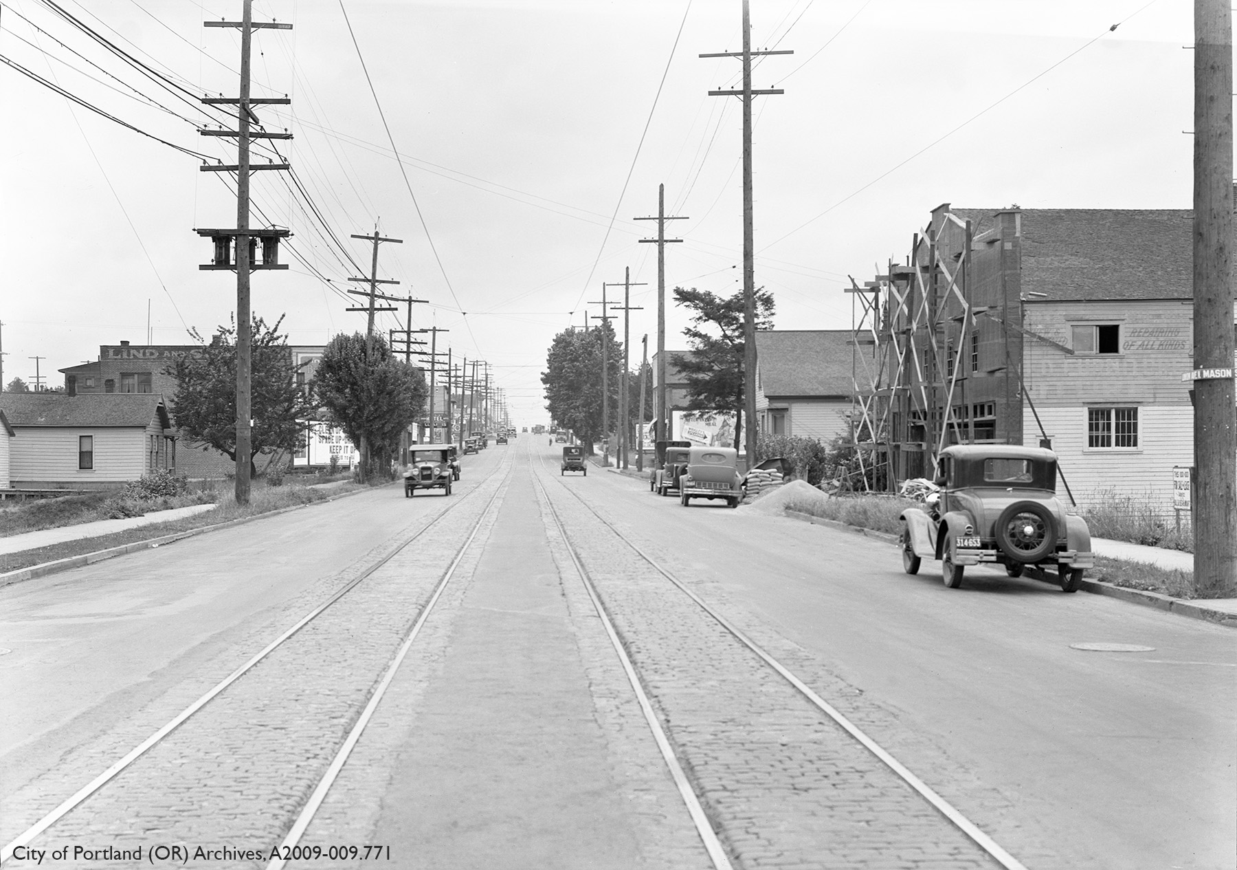 Black and white photograph of street with two sets of tracks, cars parked on the sides of the road